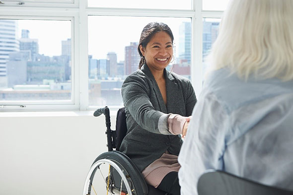 Smilende kvinne i rullestol på kontor håndhilser på en annen kvinne