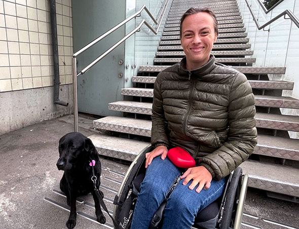 Smilende Maren Huseby i rullestol med servicehund ved siden av seg foran trapp