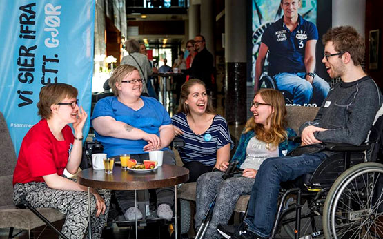 Fem smilende unge mennesker, hvorav en i rullestol, rundt kafebord