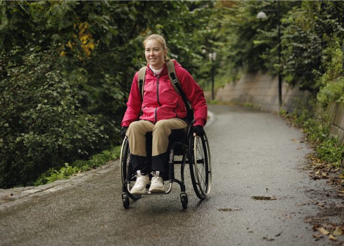 Kvinne i rullestol på gangvei omgitt av busker og trær