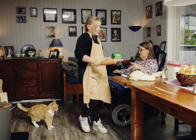 Anne Karin Johansen i rullestol instruerer barnebarn ved kjøkkenbordet med deig og kjevle 