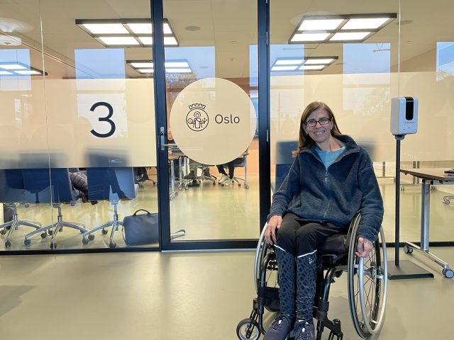 Regionstyremedlem Ingrid Njerve sitter smilende i manuell rullestol foran møterommet der den ene høringen skal foregå.