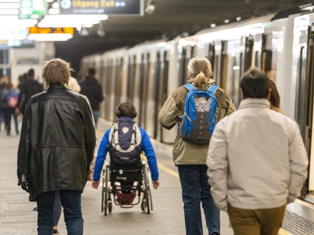 Vi ser passasjerer på en t-baneperrong i Oslo. Alle med ryggen til kamera. Noen går, en sitter i rullestol.