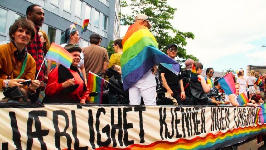Vi ser bilde fra Pride-paraden. Glade mennesker, noen i rullestol, holder regnbueflagg. Et banner med påskriften "Kjærlighet kjenner ingen grenser" er sentralt i bildet.